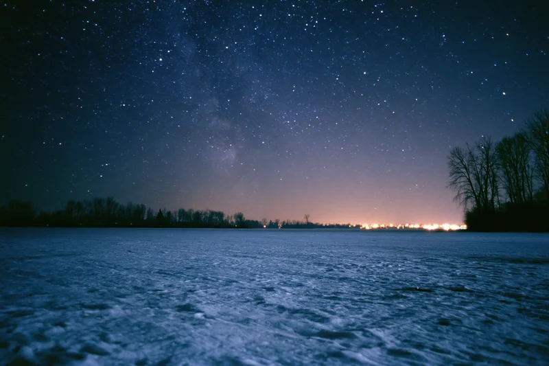 Snow-covered field under star-filled night sky with distant horizon glow