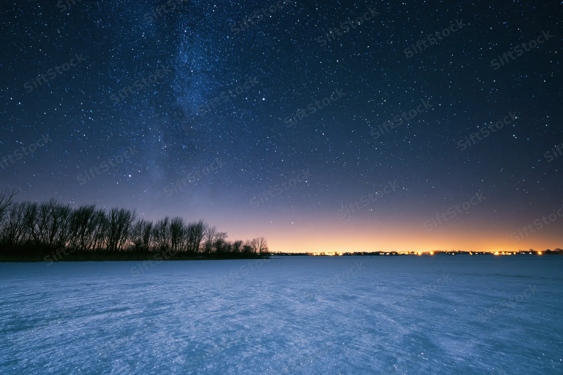 Snow-covered field under star-filled night sky with distant horizon glow