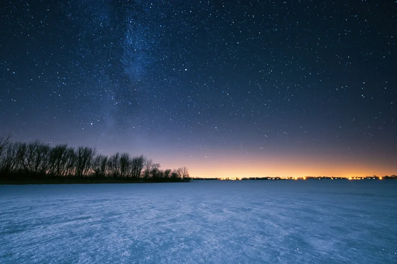 Snow-covered field under star-filled night sky with distant horizon glow
