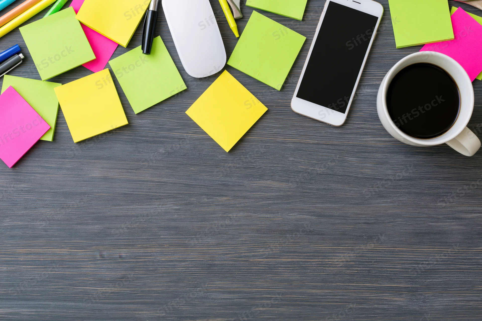 Overhead view of colorful sticky notes, smartphone, coffee mug, pens and mouse arranged along top edge of dark wooden desk with copyspace below