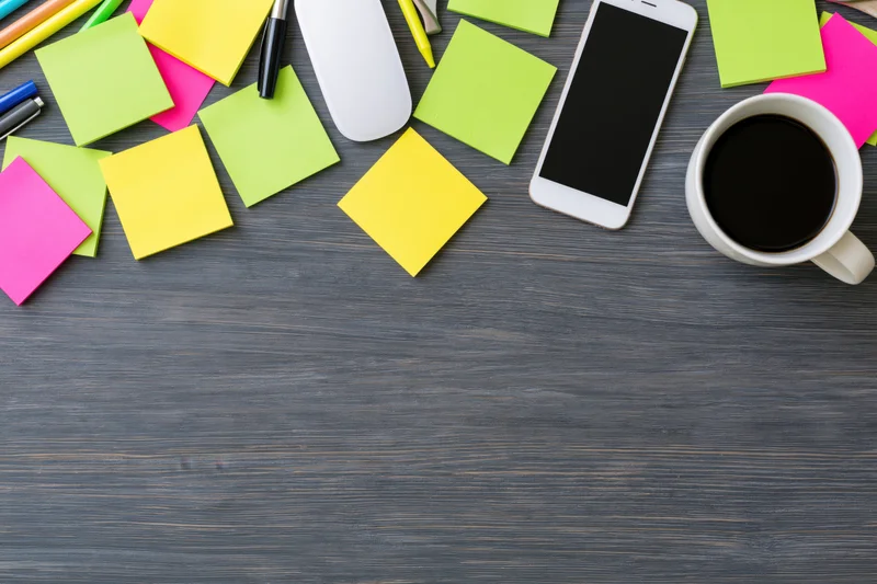 Overhead view of colorful sticky notes, smartphone, coffee mug, pens and mouse arranged along top edge of dark wooden desk with copyspace below