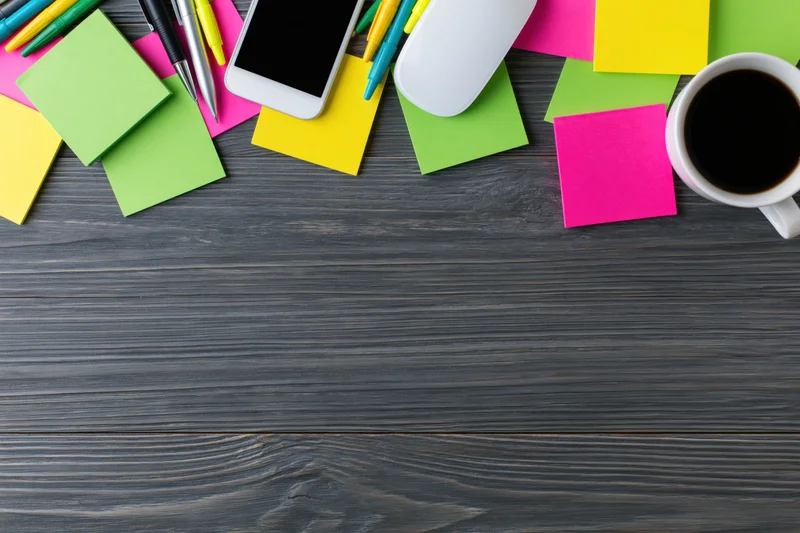 Colorful office supplies and coffee mug arranged along top edge of dark wooden desk with copyspace below