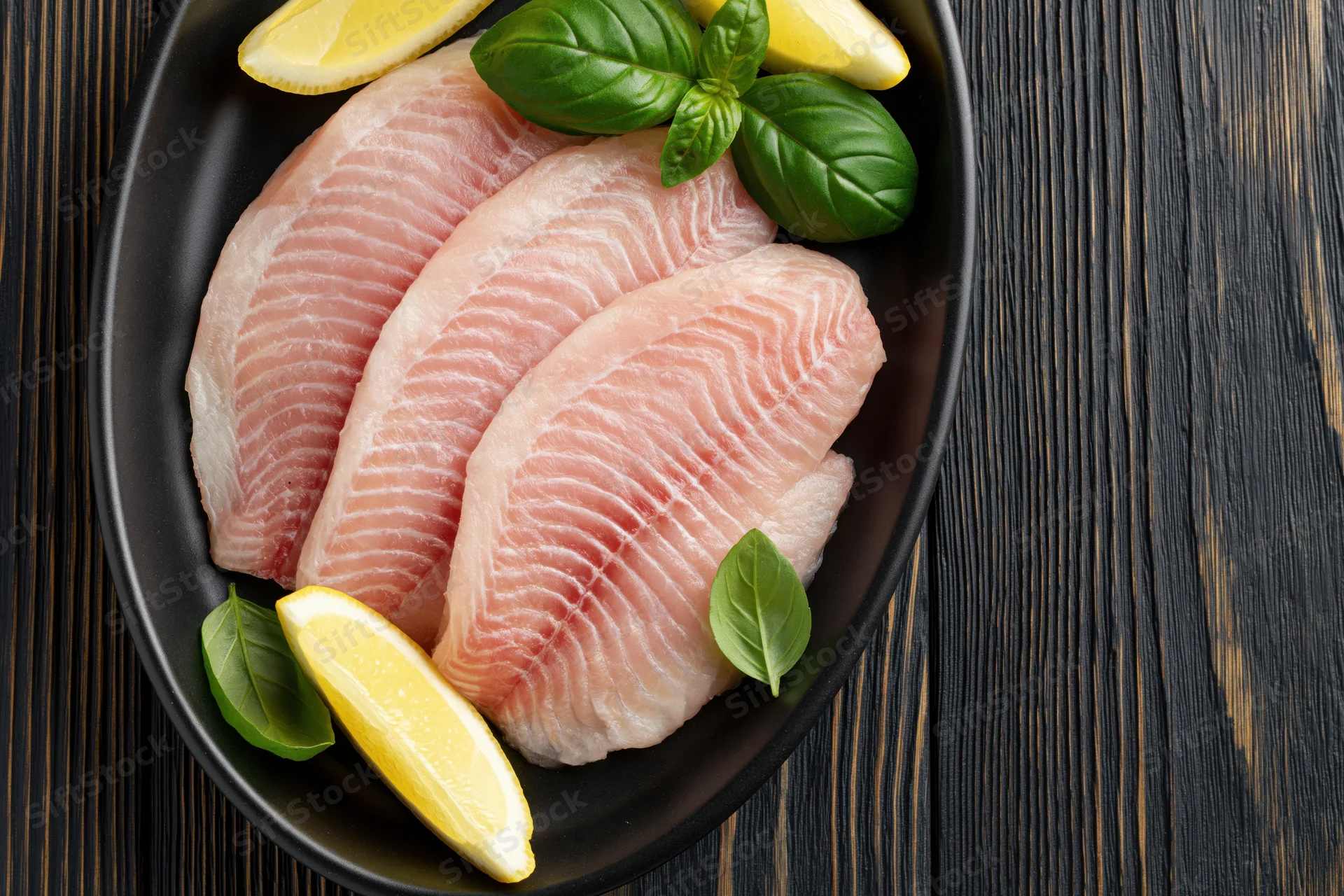 Raw tilapia fillets arranged in a black oval dish with lemon and basil garnish on dark wood surface