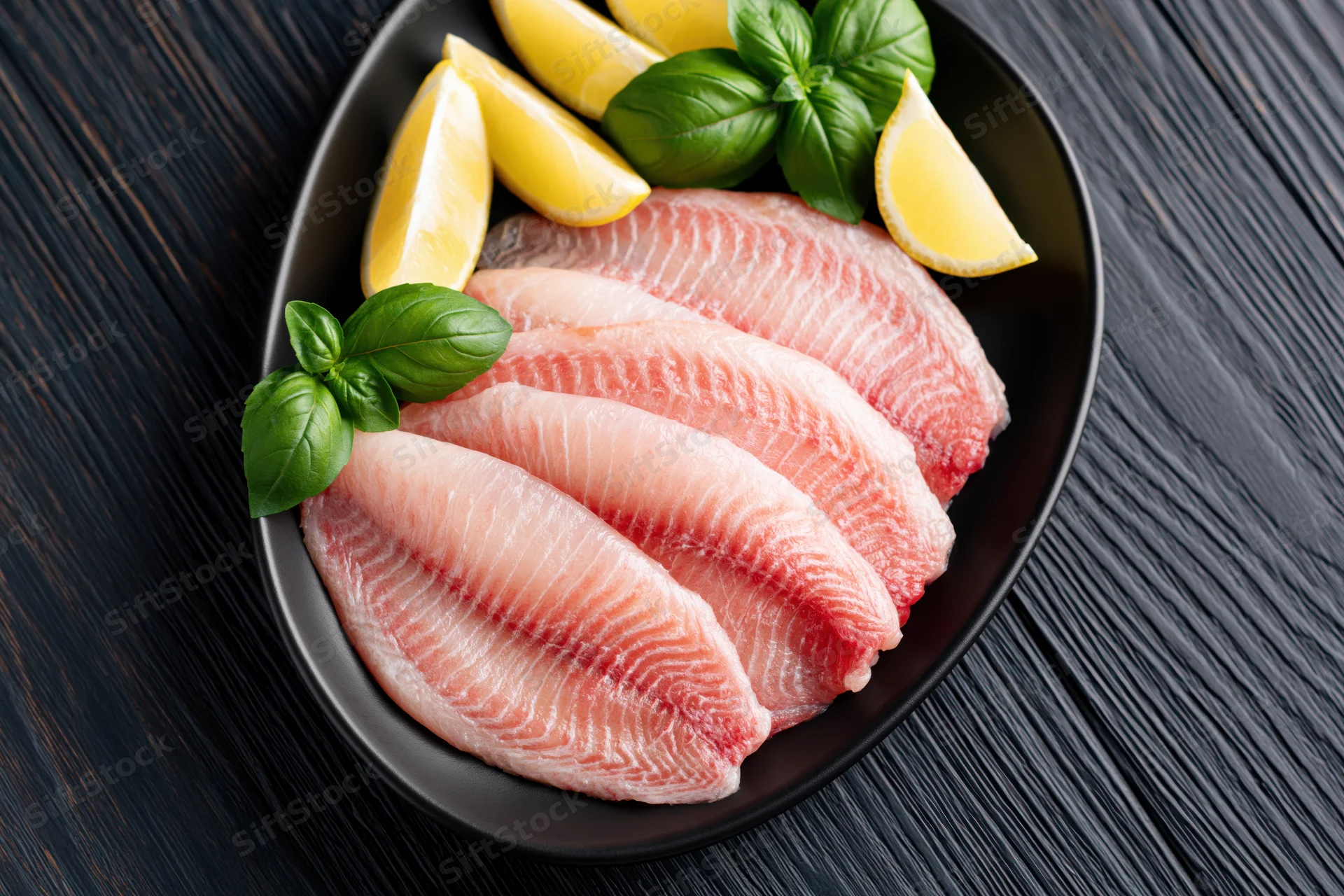 Raw tilapia fillets arranged in a black oval serving dish with lemon wedges and basil garnish on a dark wooden surface