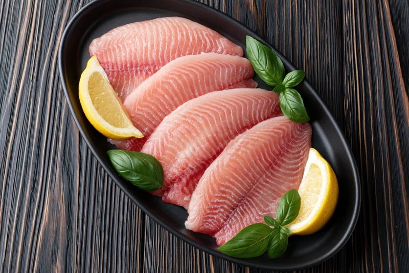 Raw tilapia fillets arranged in a black oval dish with lemon and basil on a dark wooden surface