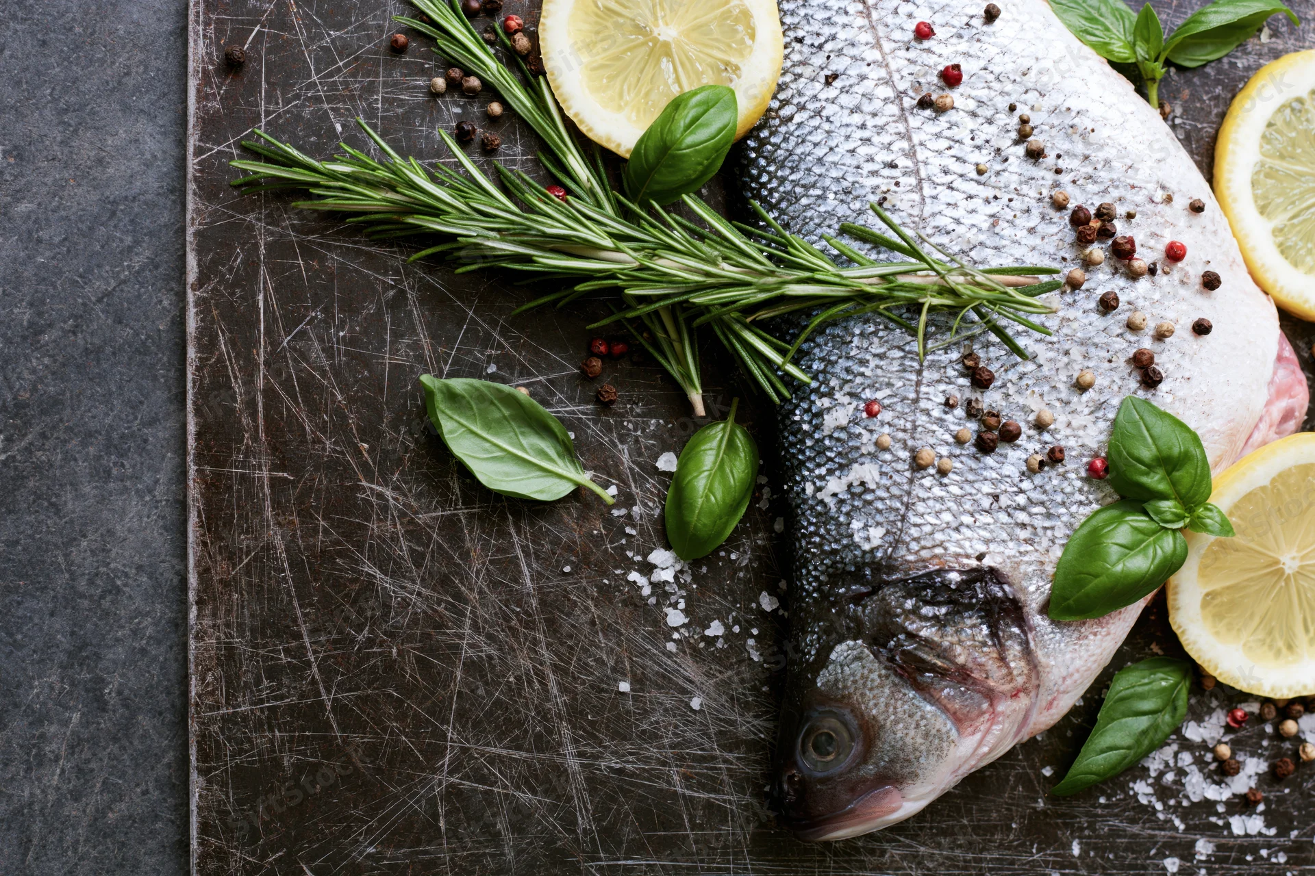 Raw whole fish on a scratched dark metal cutting board with lemon slices, rosemary, basil, pepper and salt