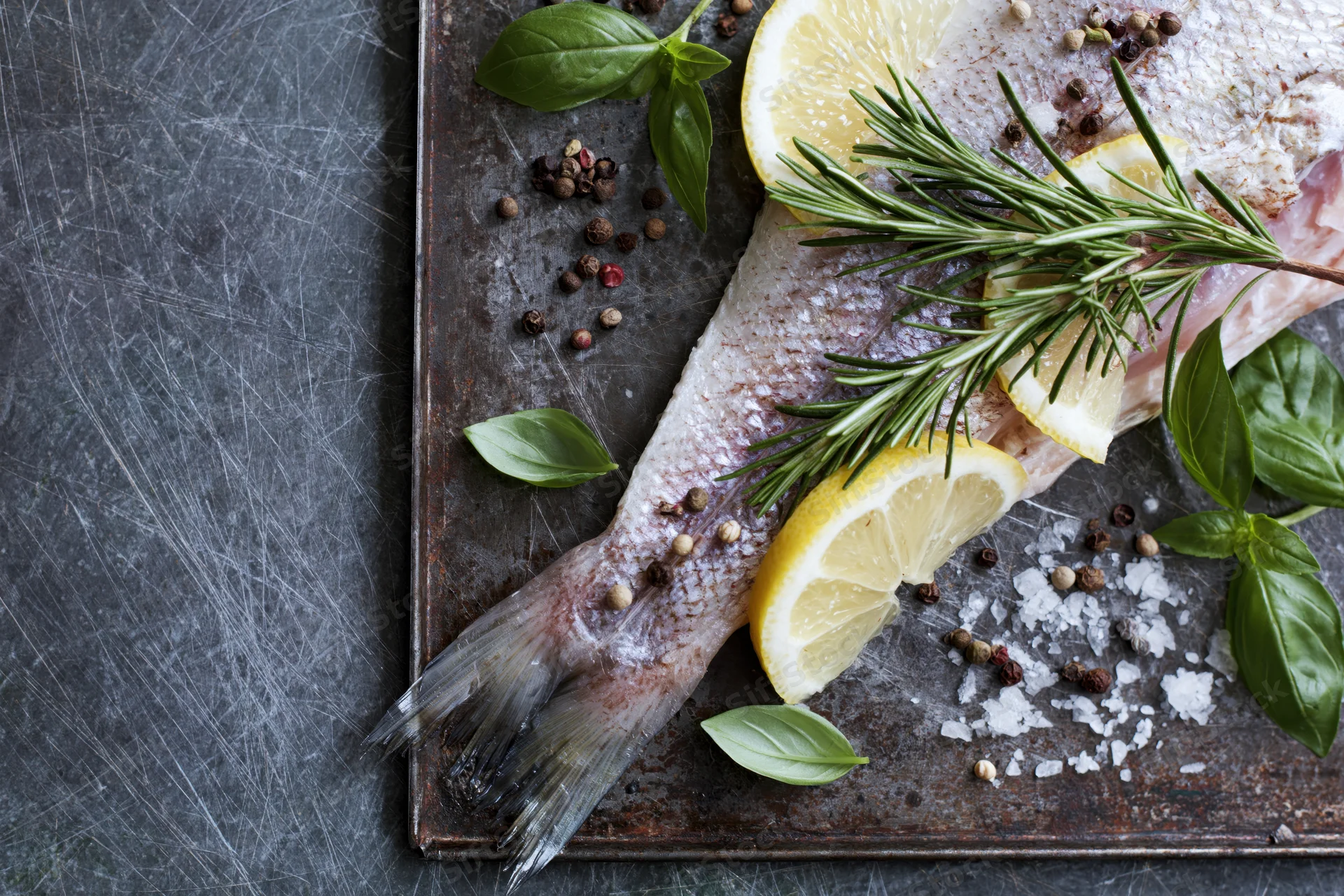 Raw whole fish with lemon, rosemary and basil on a scratched dark metal baking tray