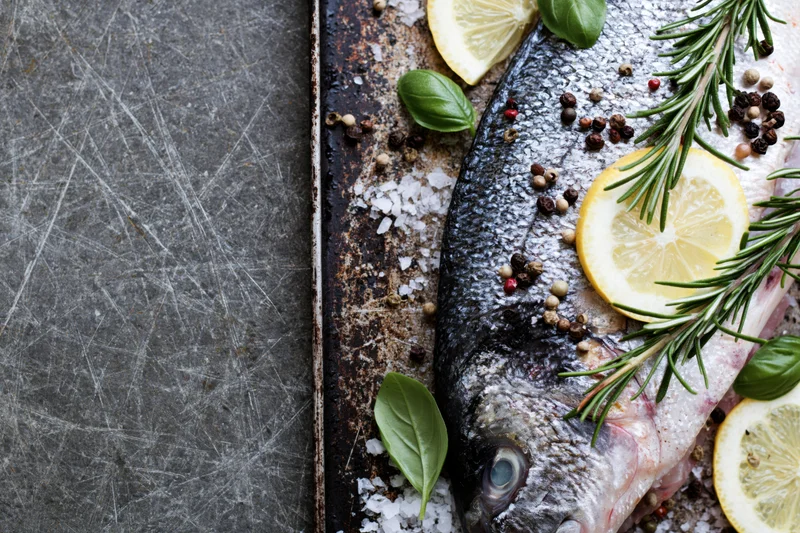 Raw whole fish on a dark scratched metal baking tray with lemon slices, rosemary, basil, and peppercorns