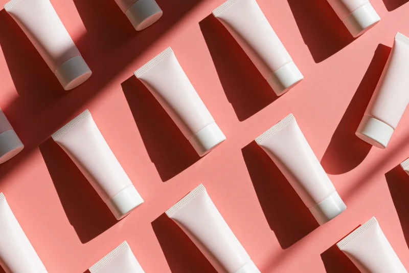 Repeating pattern of blank white cosmetic cream tubes arranged on pink surface in flat lay overhead view