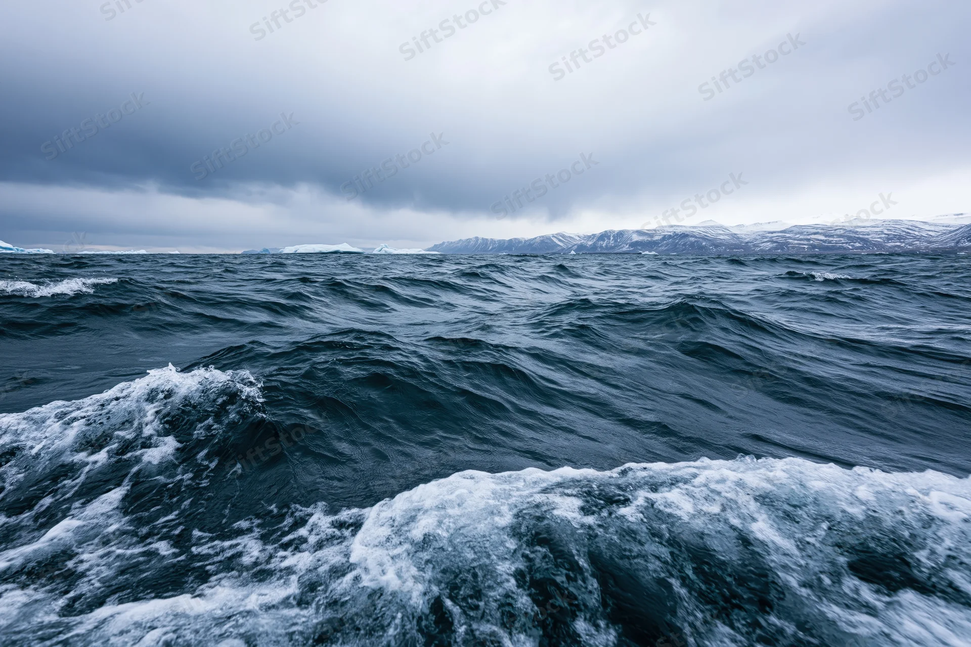 Stormy ocean waves churning under dark clouds with snowy mountains in the distance