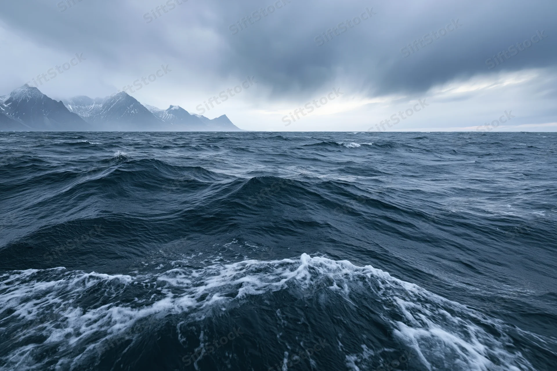 Rough dark ocean waves under stormy sky with snow-capped mountains in distance