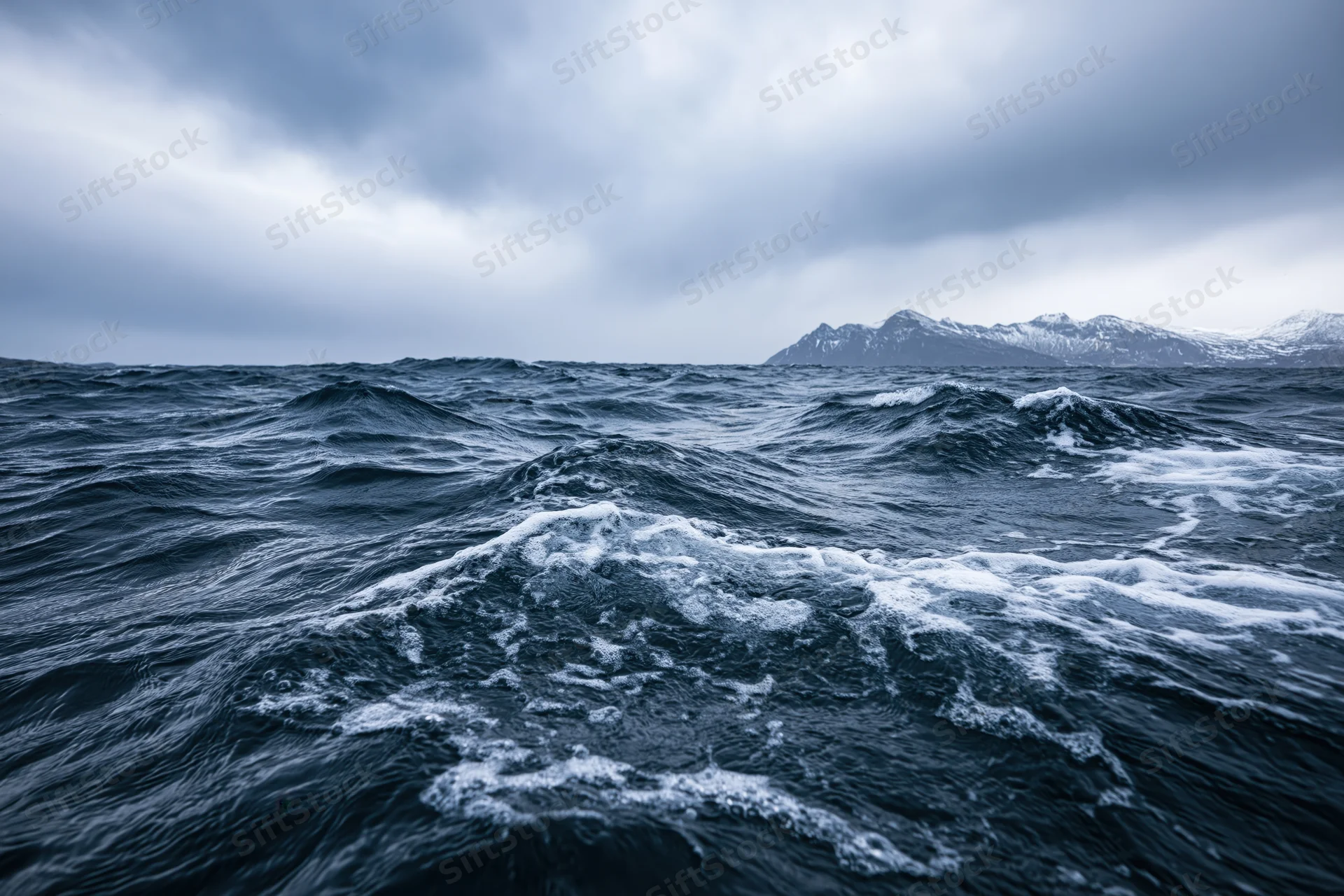 Rough dark ocean waves under stormy sky with snow-capped mountains in distance