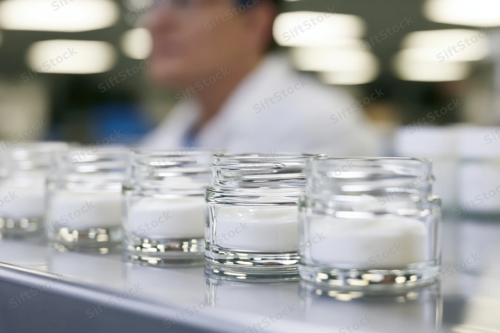 Row of open glass jars filled with white cream on a conveyor in a laboratory setting