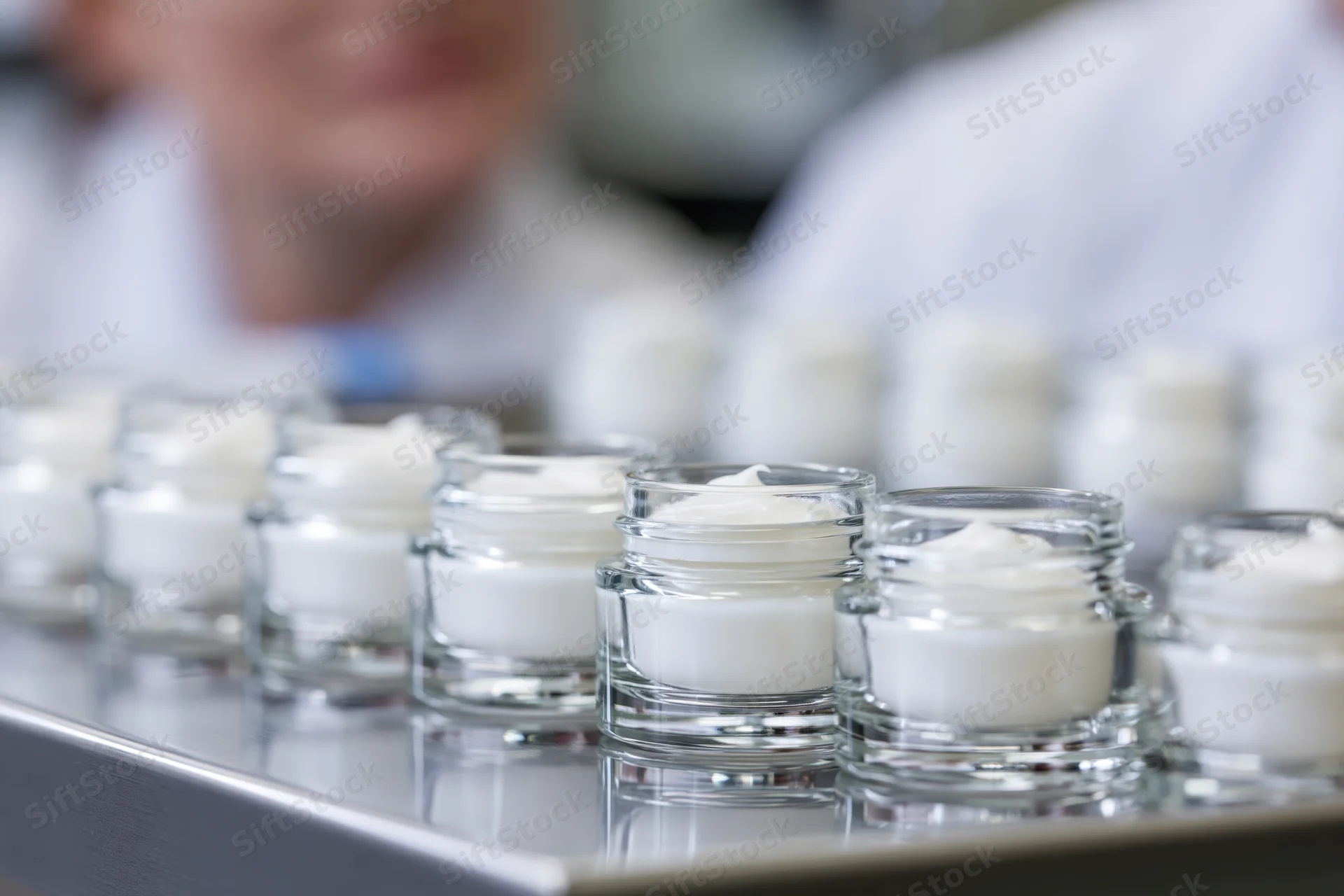 Row of small open glass jars filled with white cream arranged on a metal tray in a laboratory setting