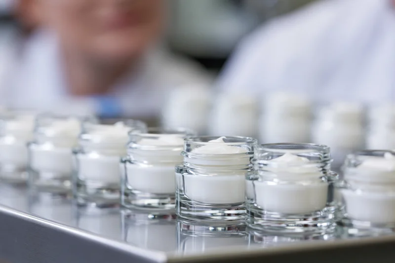 Row of small open glass jars filled with white cream arranged on a metal tray in a laboratory setting