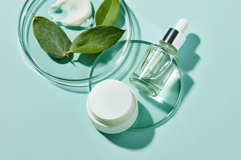 Skincare flatlay with glass dropper bottle, white cream jar, and green leaves on teal background