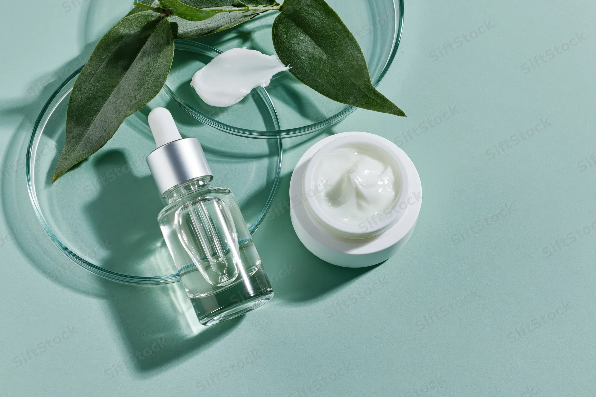 Glass dropper serum bottle and white cream jar with green leaves on mint background