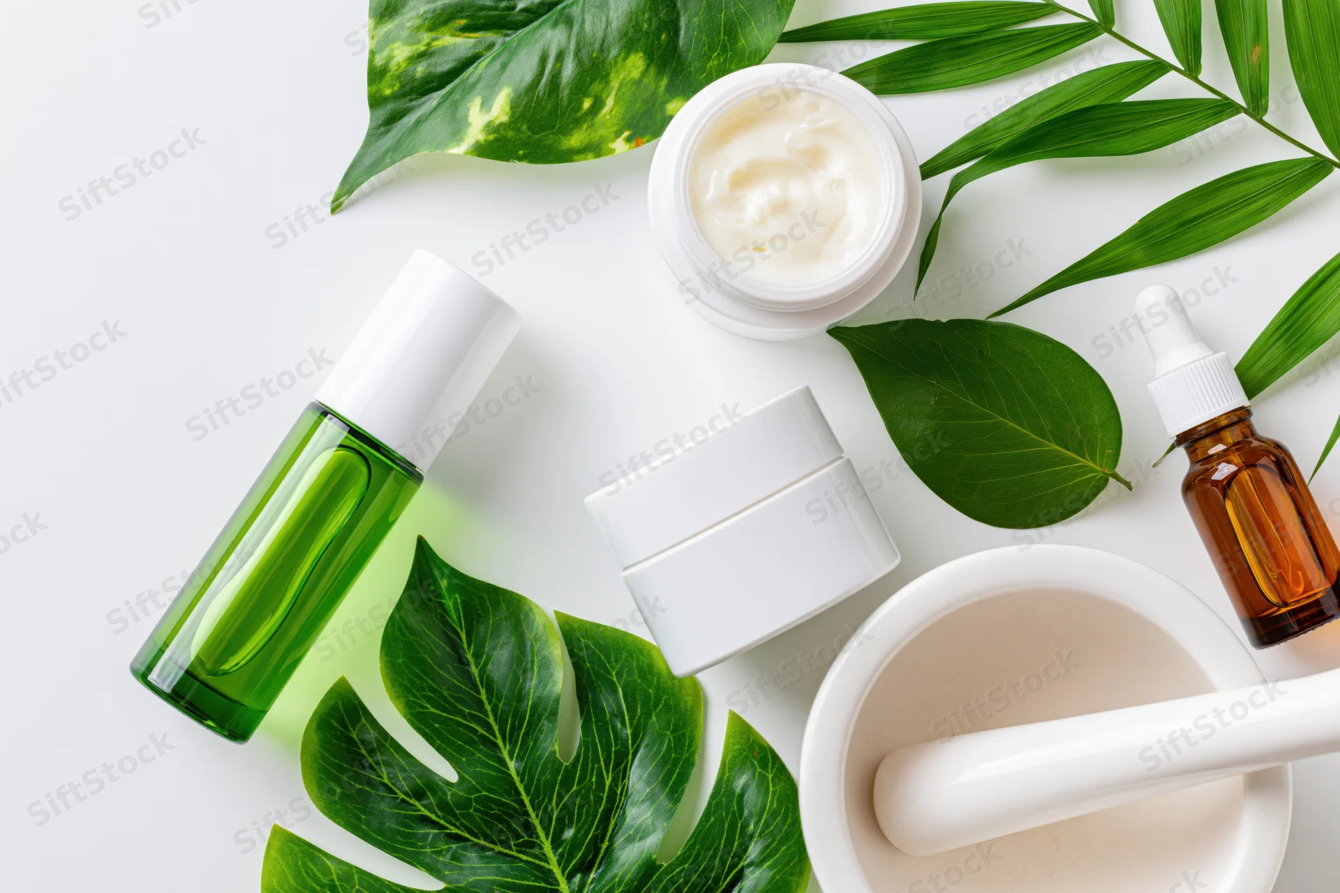 Skincare products and green leaves arranged on white surface with mortar and pestle