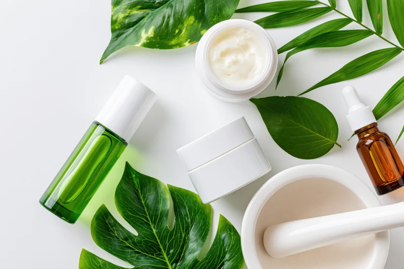 Skincare products and green leaves arranged on white surface with mortar and pestle