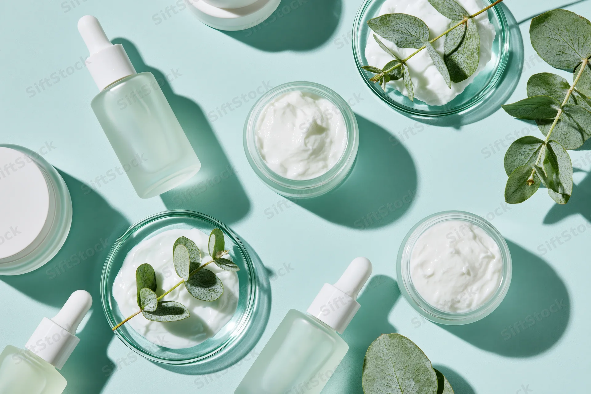 Skincare products arranged in flat lay on mint blue surface with eucalyptus leaves