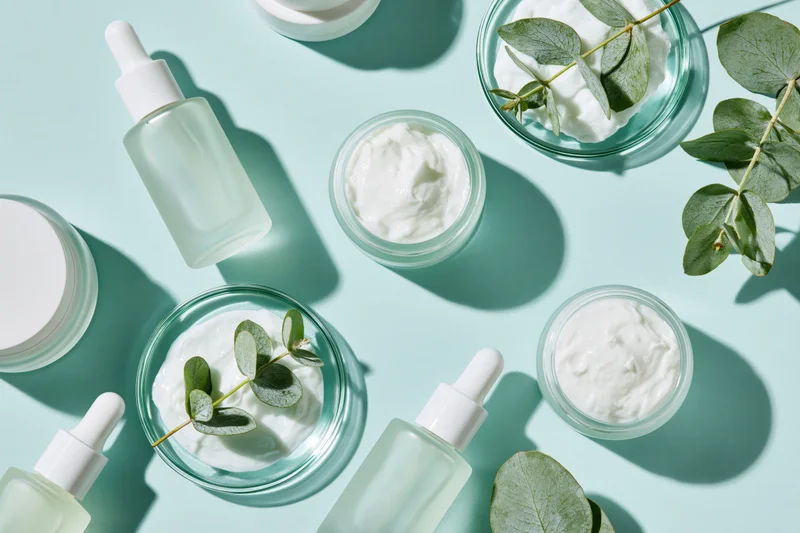 Skincare products arranged in flat lay on mint blue surface with eucalyptus leaves