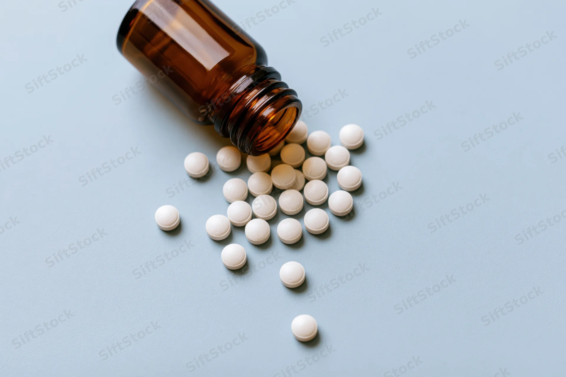 Small amber glass bottle lying on its side with white pills spilled onto a light blue surface
