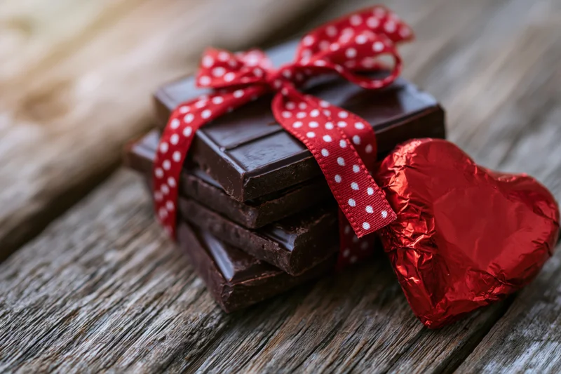 Stack of dark chocolate squares tied with red polka-dot ribbon beside a foil-wrapped heart on wooden surface