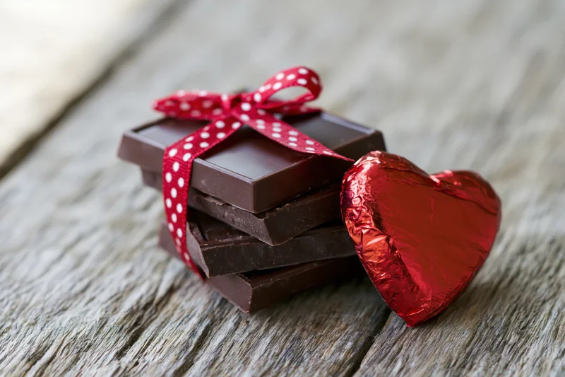 Stack of dark chocolate squares tied with red polka-dot ribbon beside a red foil-wrapped heart on wooden surface