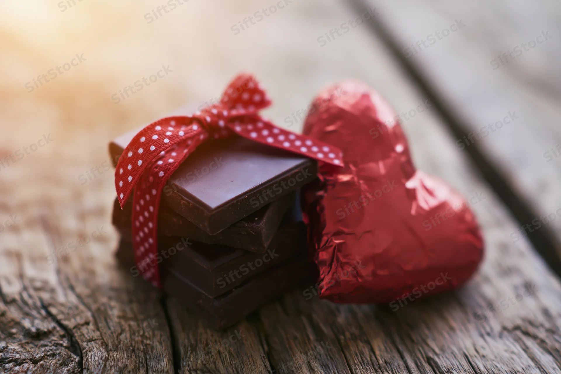 Stack of dark chocolate squares tied with red polka-dot ribbon beside a heart-shaped foil-wrapped candy on a rustic wooden surface
