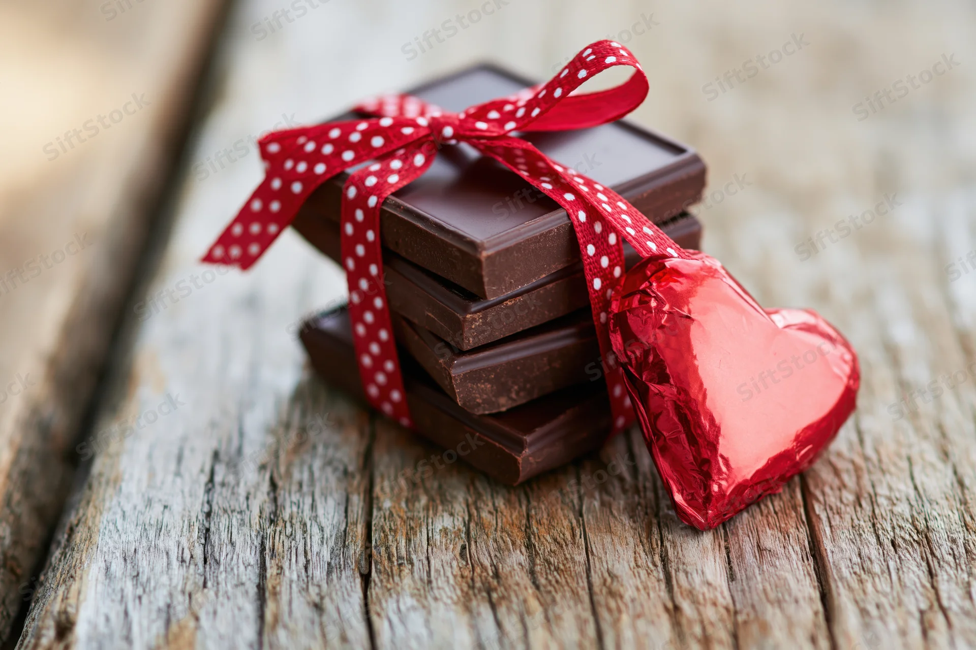 Stack of dark chocolate bars tied with red polka-dot ribbon and heart-shaped foil candy on rustic wooden surface