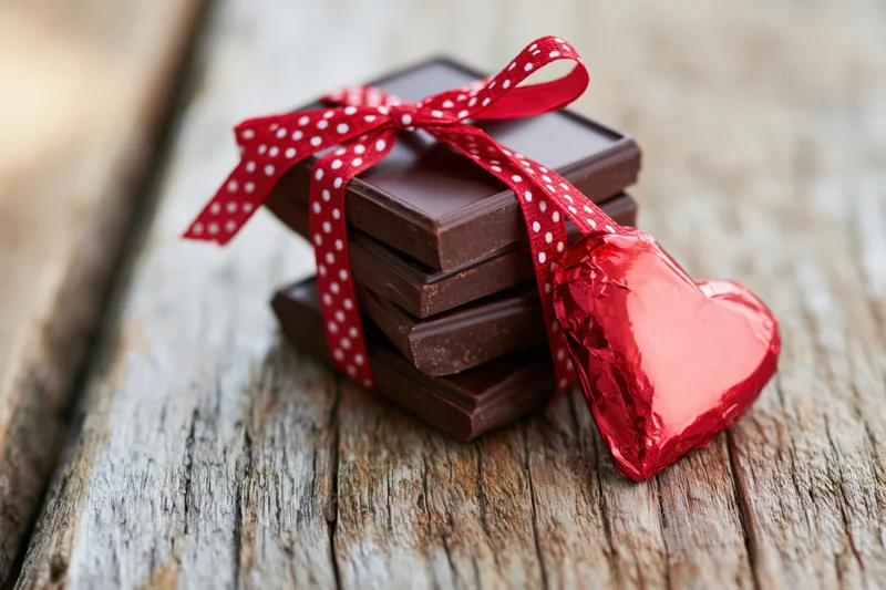 Stack of dark chocolate bars tied with red polka-dot ribbon and heart-shaped foil candy on rustic wooden surface