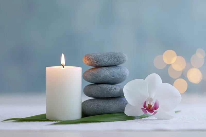 Lit white candle beside stacked grey zen stones and white orchid on textured surface with soft bokeh lights in background