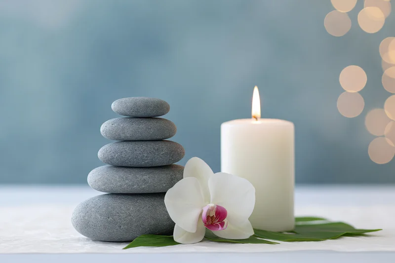 Stacked smooth grey zen stones beside a lit white pillar candle on a white surface with green leaves and a white orchid flower