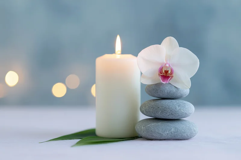 Lit white candle beside stacked grey zen stones with white orchid on top on a table with soft bokeh lights in background