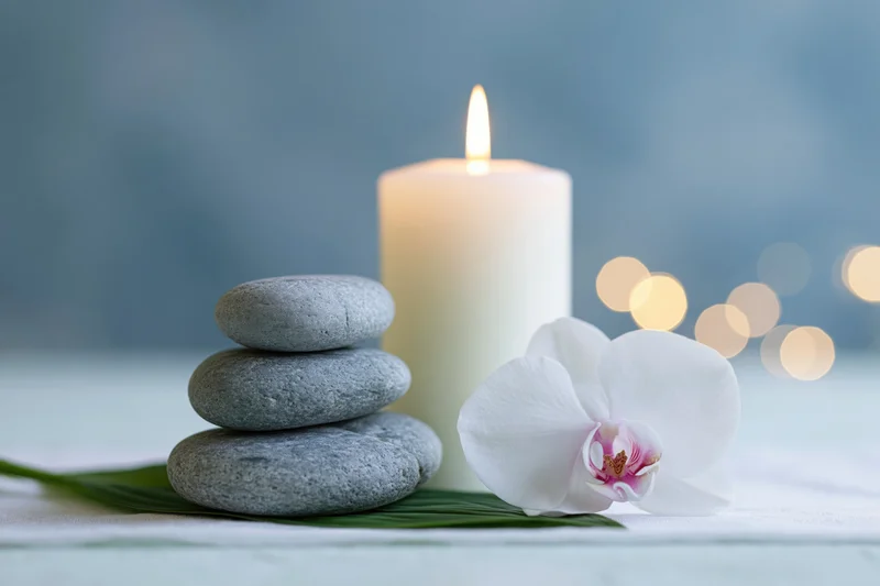 Stacked smooth grey zen stones beside a lit white pillar candle and a white orchid on a towel with bokeh lights in the background