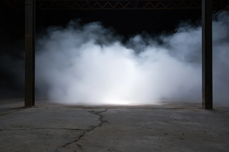 Thick white smoke rising from cracked concrete floor between metal beams in dark industrial warehouse interior