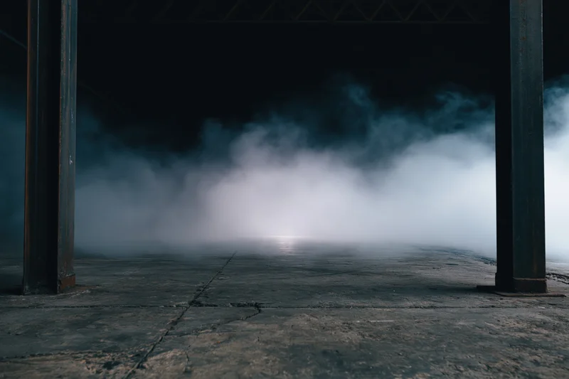 Thick smoke rising from a cracked concrete floor between steel pillars in a dark industrial warehouse interior