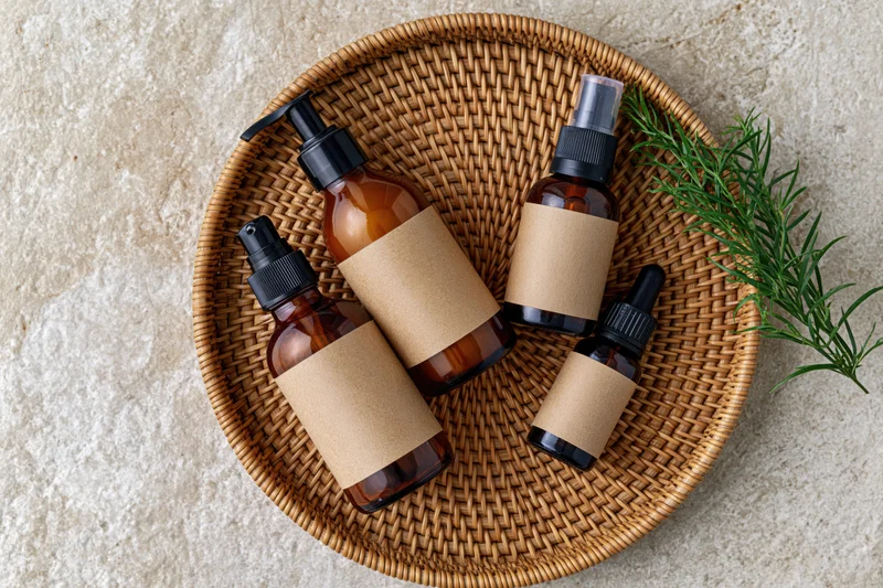 Amber glass skincare bottles with blank kraft paper labels arranged on a woven wicker basket with a rosemary sprig on a stone surface