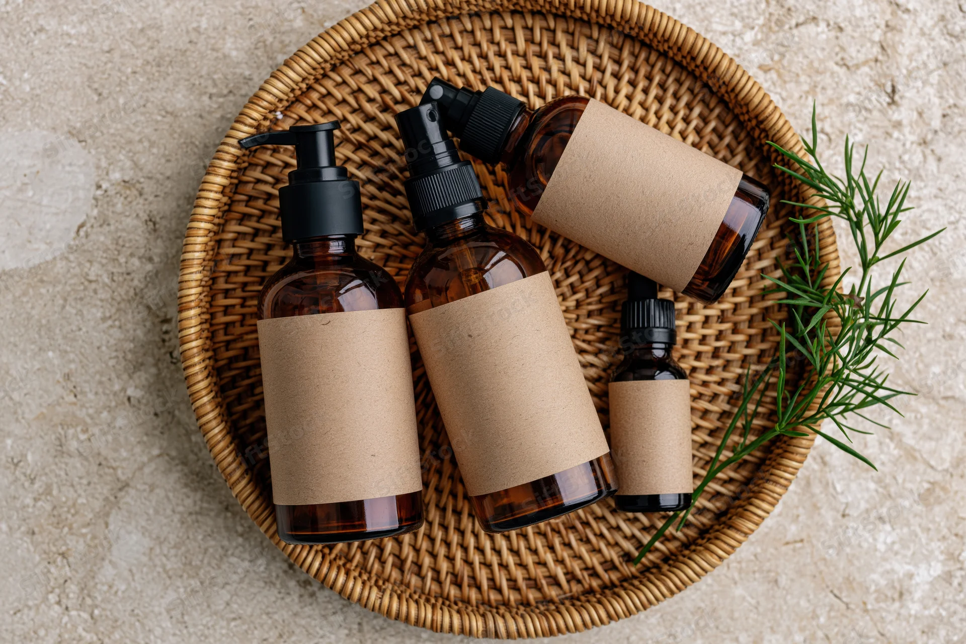 Amber glass skincare bottles with blank kraft paper labels arranged on a woven wicker tray with rosemary sprig