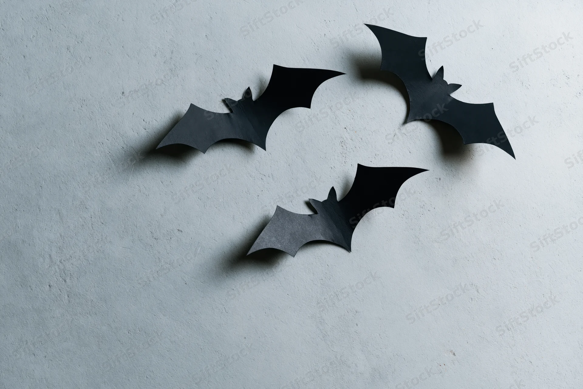 Three black paper bat cutouts arranged in a loose flying cluster on a textured gray wall