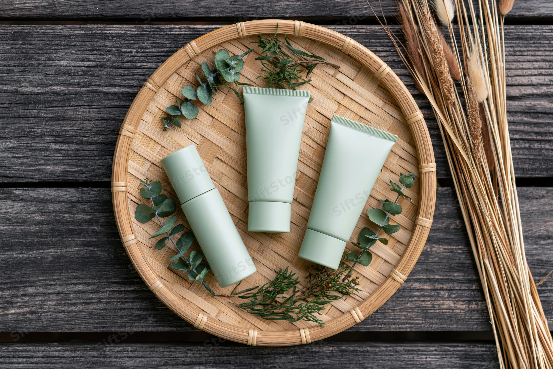 Three matte sage-green skincare tubes and a slim serum bottle arranged on a woven bamboo tray with greenery and dried wheat on a dark wooden surface