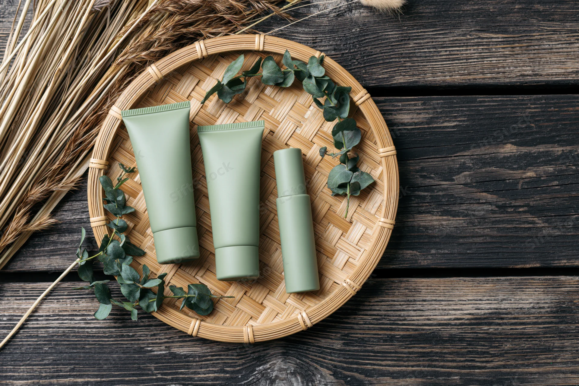 Three matte sage-green skincare tubes and a slim serum bottle arranged on a woven bamboo tray with eucalyptus and dried grass on dark wooden surface
