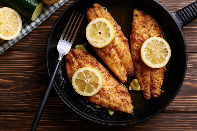 Three pan-seared fish fillets topped with lemon slices in a black skillet on a wooden table with a fork beside it