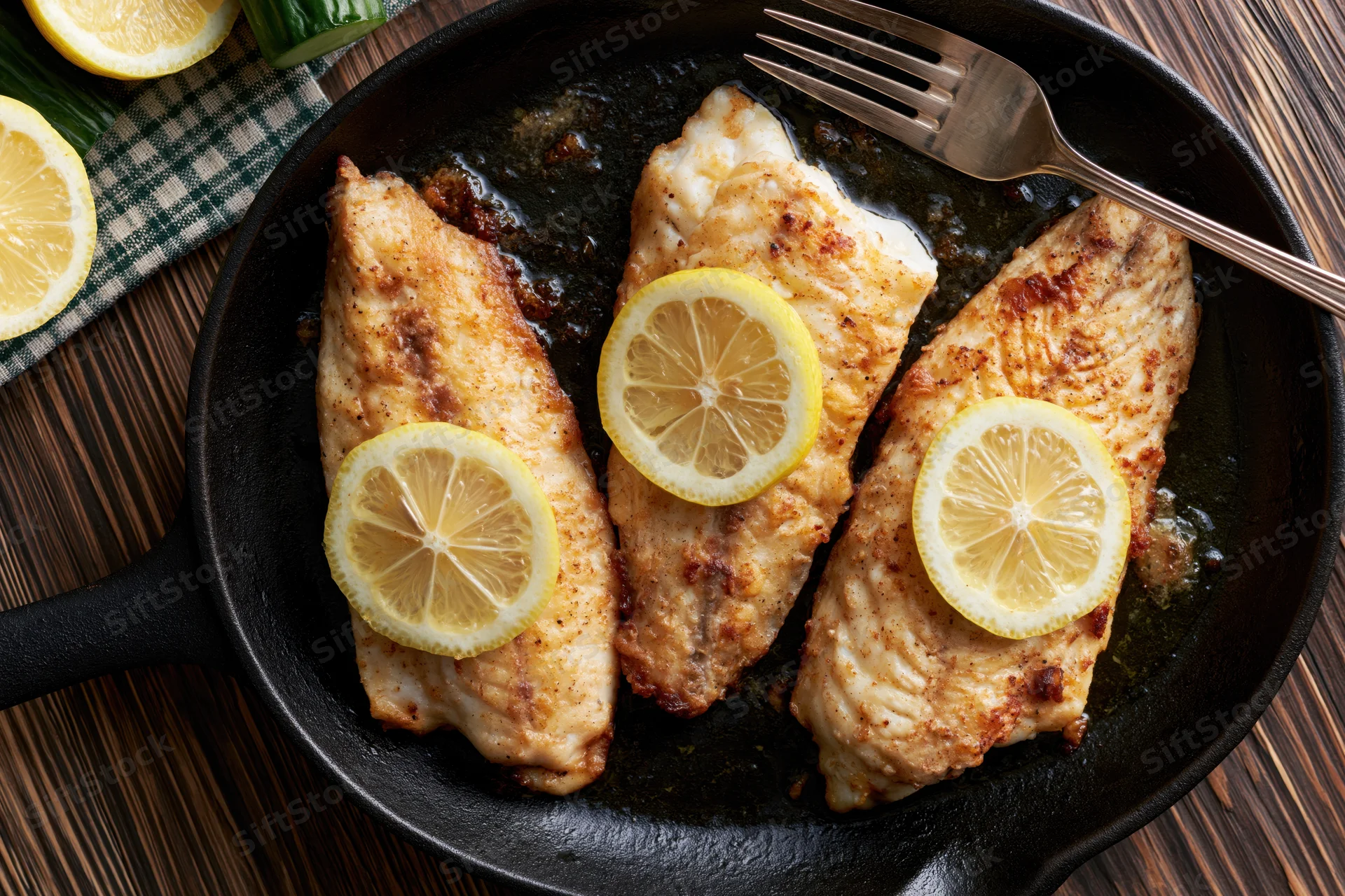 Three pan-seared fish fillets topped with lemon slices in a cast iron skillet on a wooden table