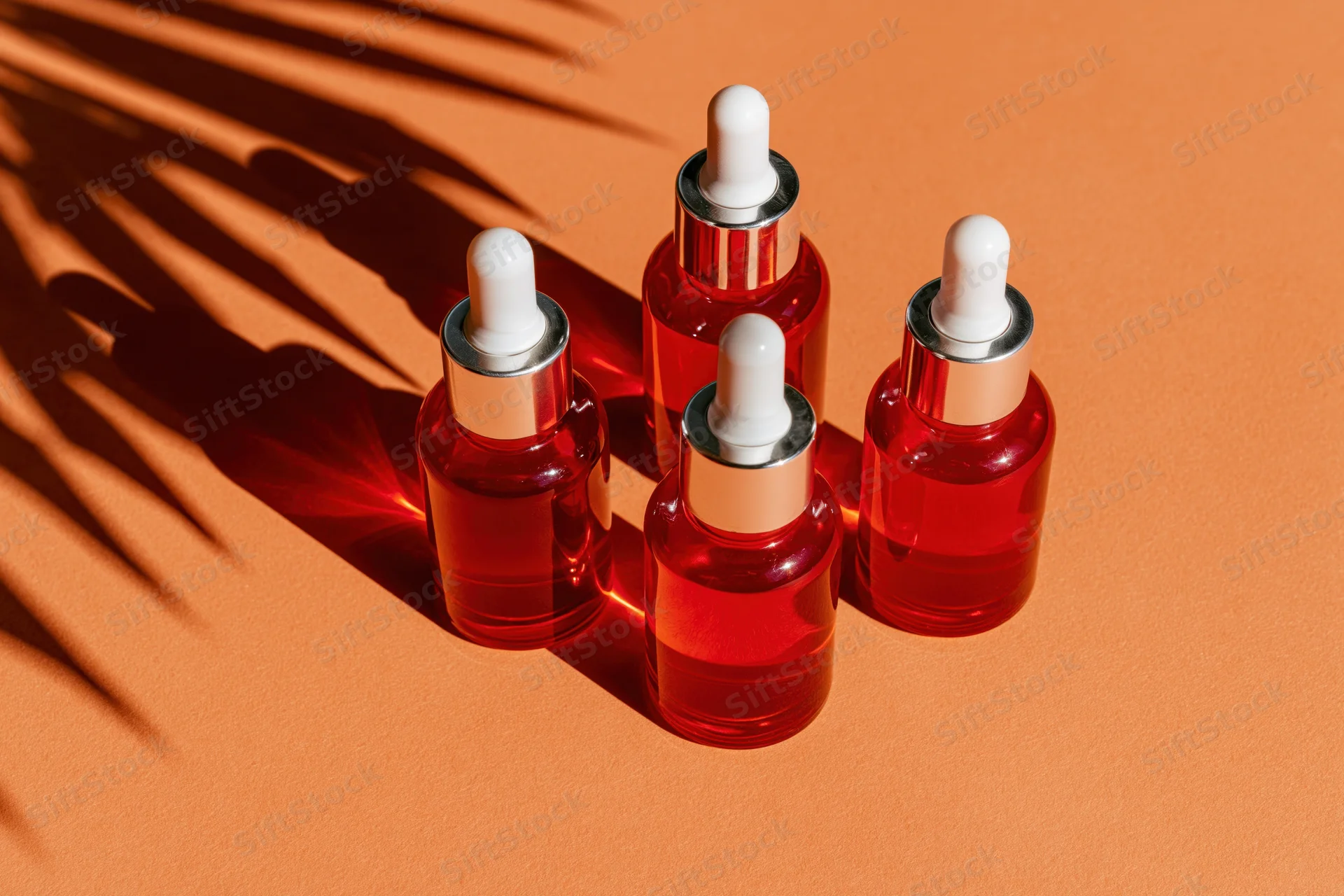 Four red glass dropper bottles with white caps arranged on orange surface with palm shadow