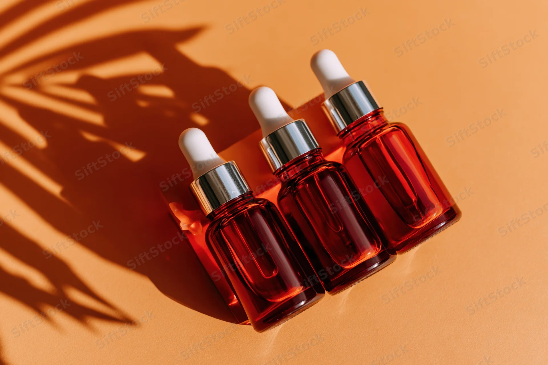 Three amber glass dropper bottles arranged in a row on a warm terracotta surface with palm leaf shadows