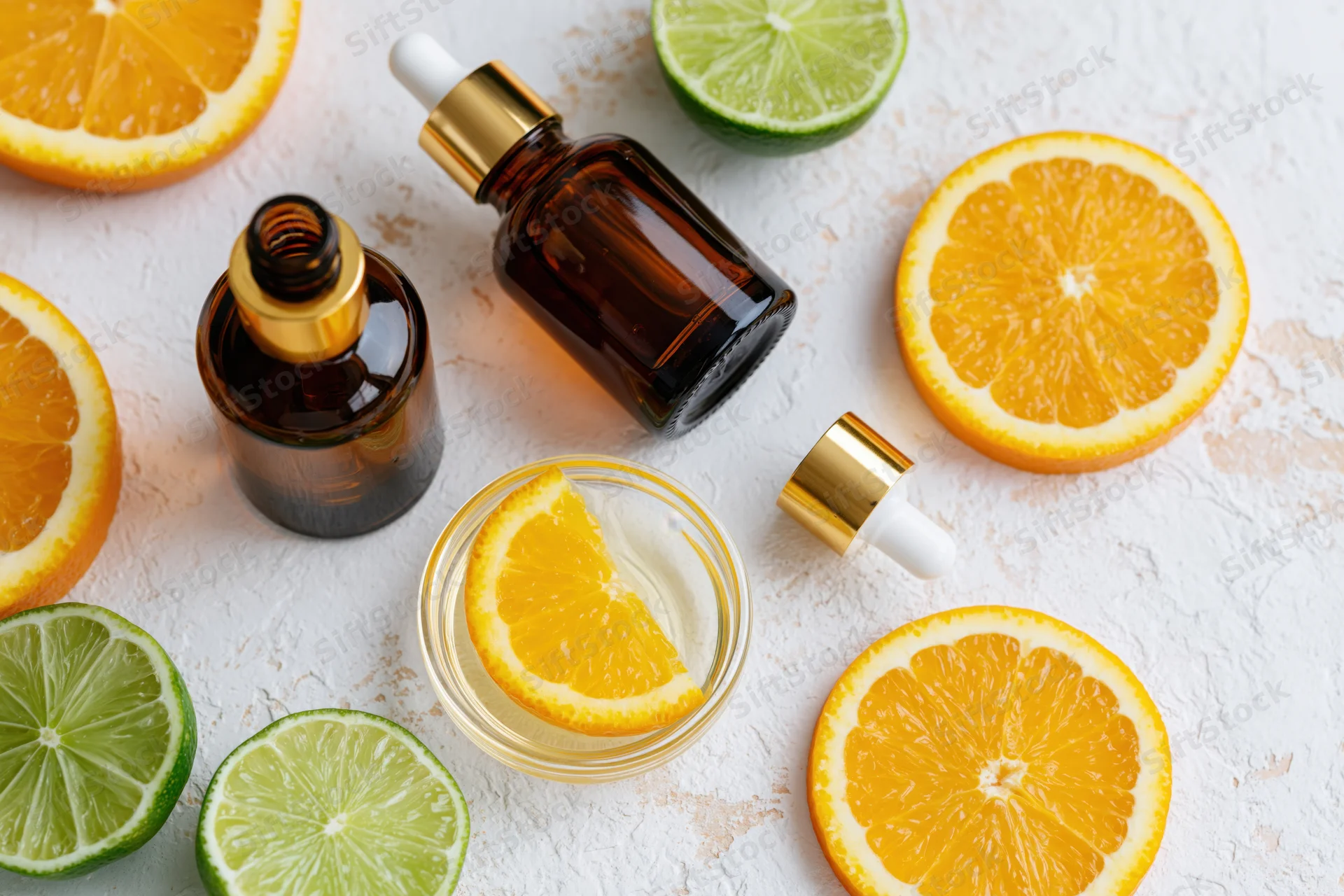 Sliced oranges and limes with amber glass dropper bottles and a jar of cream on a white textured surface
