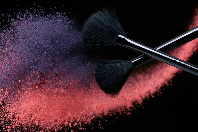 Two black makeup brushes mid-swipe releasing red and purple powder against black background