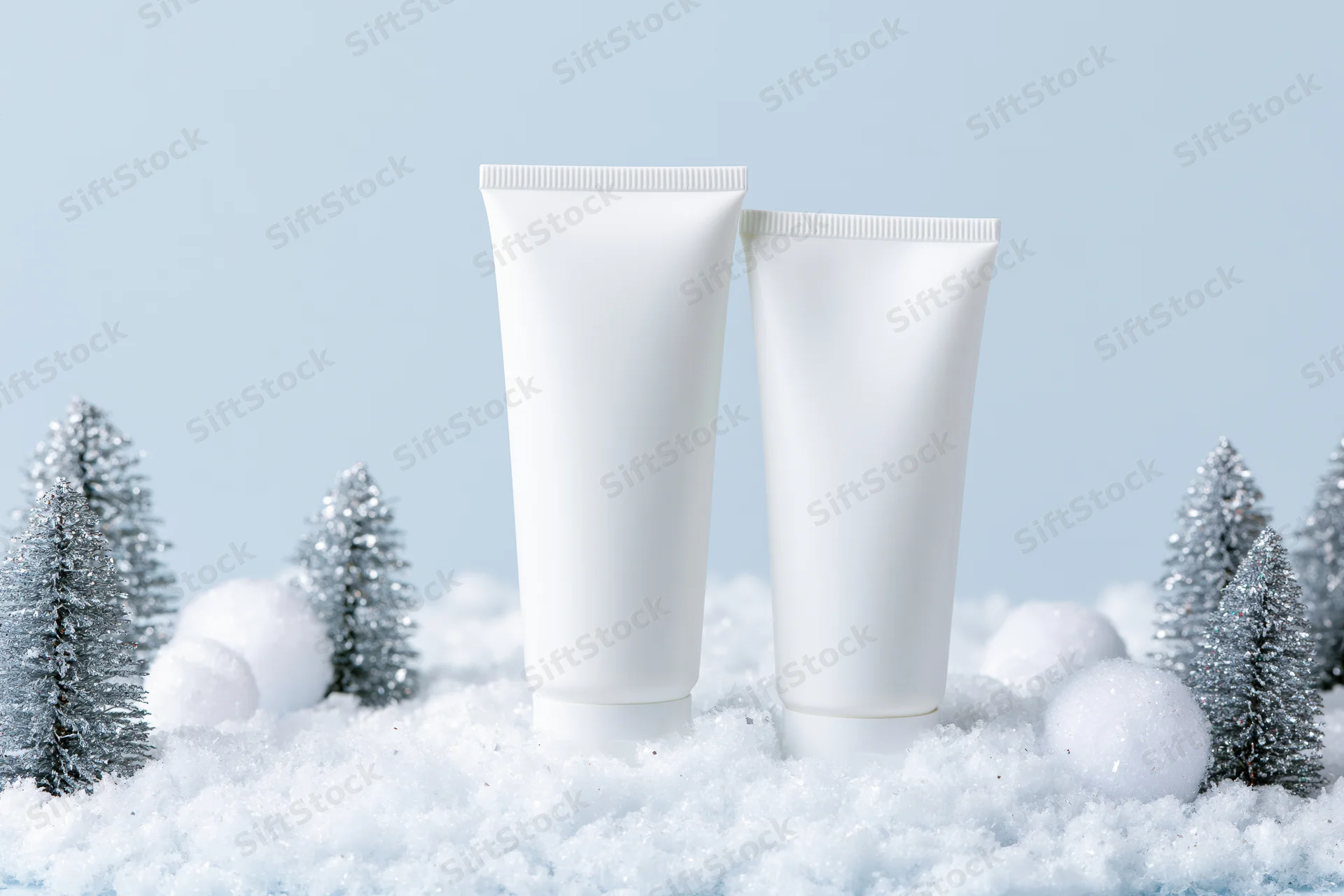 Two blank white cosmetic tubes standing on snow with miniature silver trees and snowballs around them