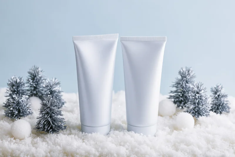 Two blank white cosmetic tubes standing on artificial snow with miniature silver trees and white pom poms around them